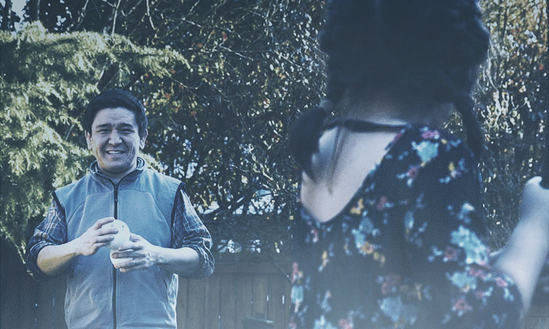 A man in a gray fleece vest holding a wiffle ball smiles at his daughter who is in the foreground poised and ready to swing with a bat.