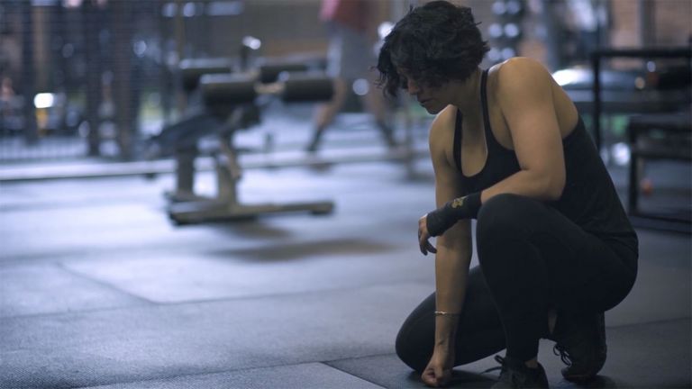 Zee Wolters in black tank top and workout tights kneels on one knee as she takes a break from lifting weights at the gym.